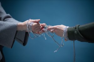 Close-up of two hands wrapped in silver chains, symbolizing connection or struggle, set against a blue backdrop.